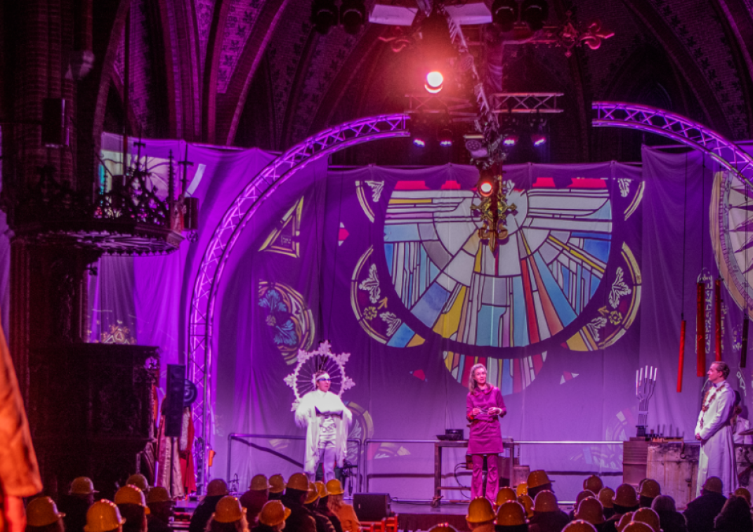 Sfeerfoto van theatervoorstelling 'Nors Staal' in Sappemeer. Er staan drie mensen op het toneel in paars licht voor glas-in-lood ramen. Achterin het publiek kijkt een man naar het podium.