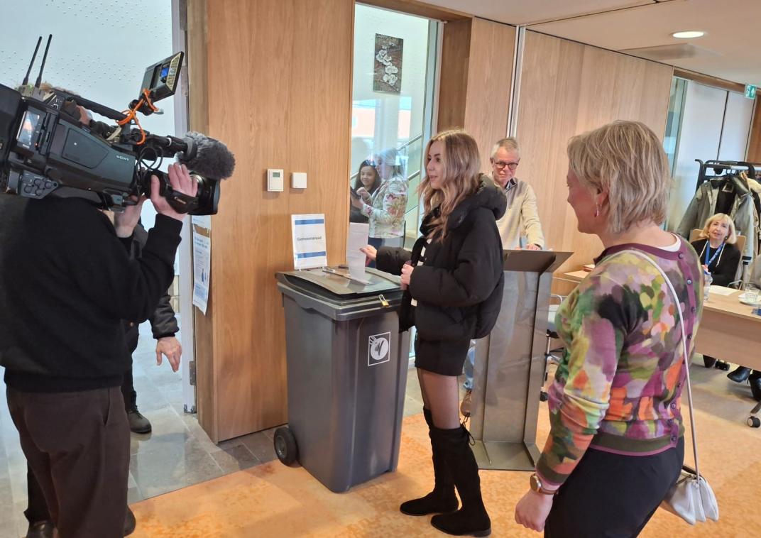 Burgemeester Erica van Lente brengt samen met Nicky (18) haar stem uit. Nicky mag dit jaar voor het eerst stemmen. Ze staan beide in de raadzaal van het Huis van Cultuur en Bestuur. Een cameraman filmt hen.