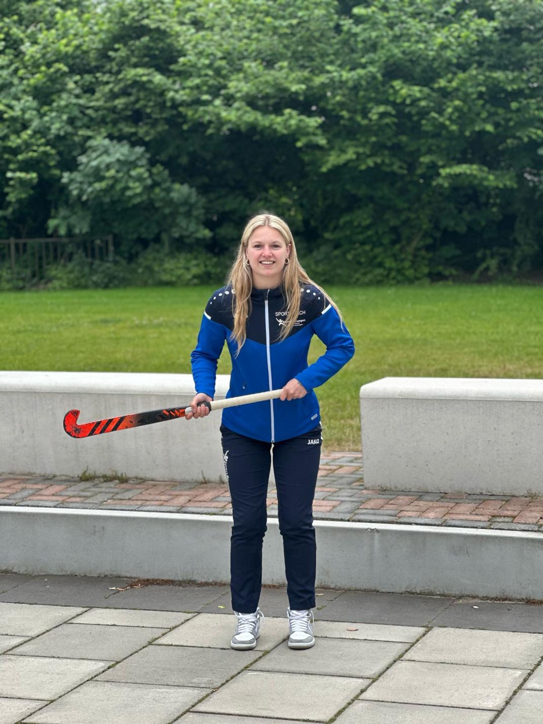 Lachende foto van beweegcoach Claudia met een hockeystick.