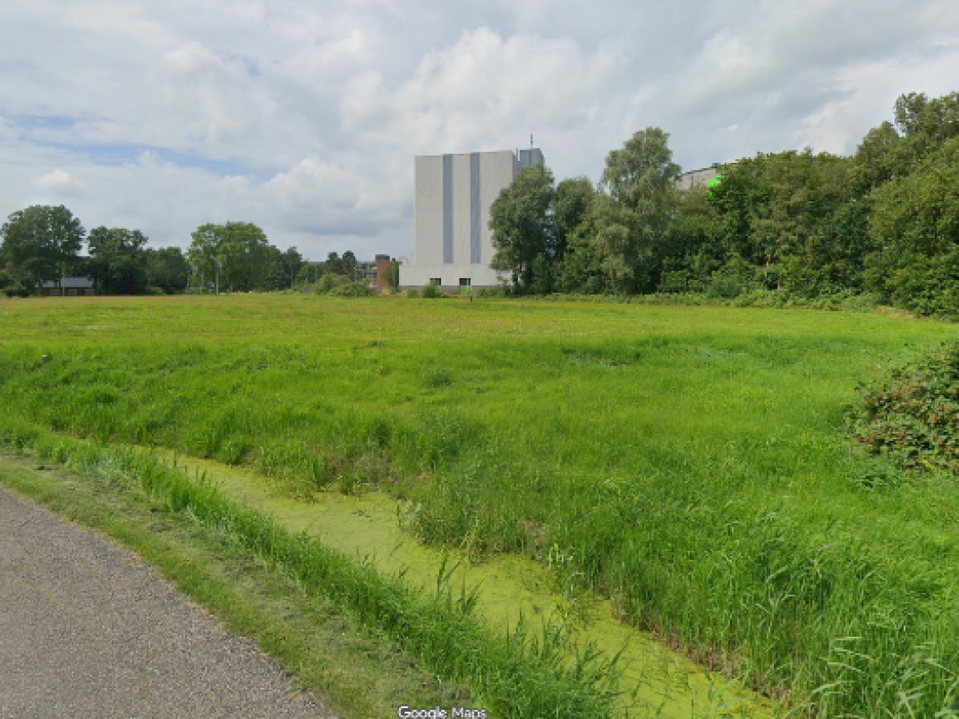 Straatbeeld van een open grasveld met sloot langs de weg, bomen aan de rand en een groot industrieel gebouw op de achtergrond.