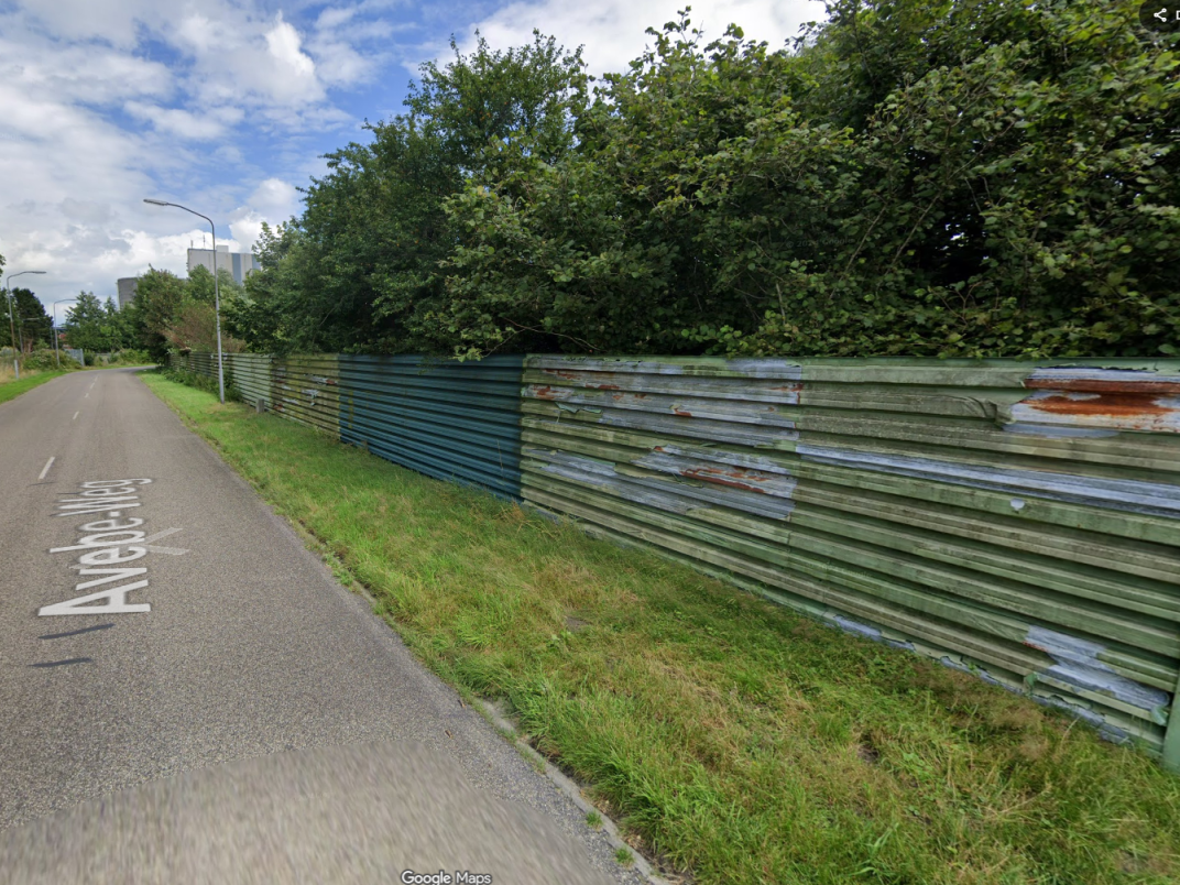 Straatbeeld van een weg met aan de rechterzijde een groen metalen afscheiding en dichte beplanting, met bebouwing op de achtergrond.