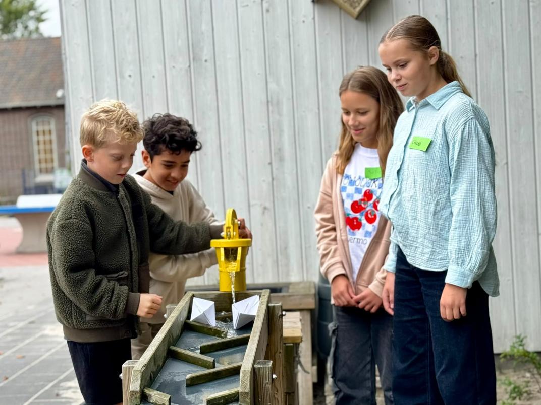 Vier kinderen zijn aan het spelen met een waterbaan. Ze laten hier papieren bootjes vanaf glijden.