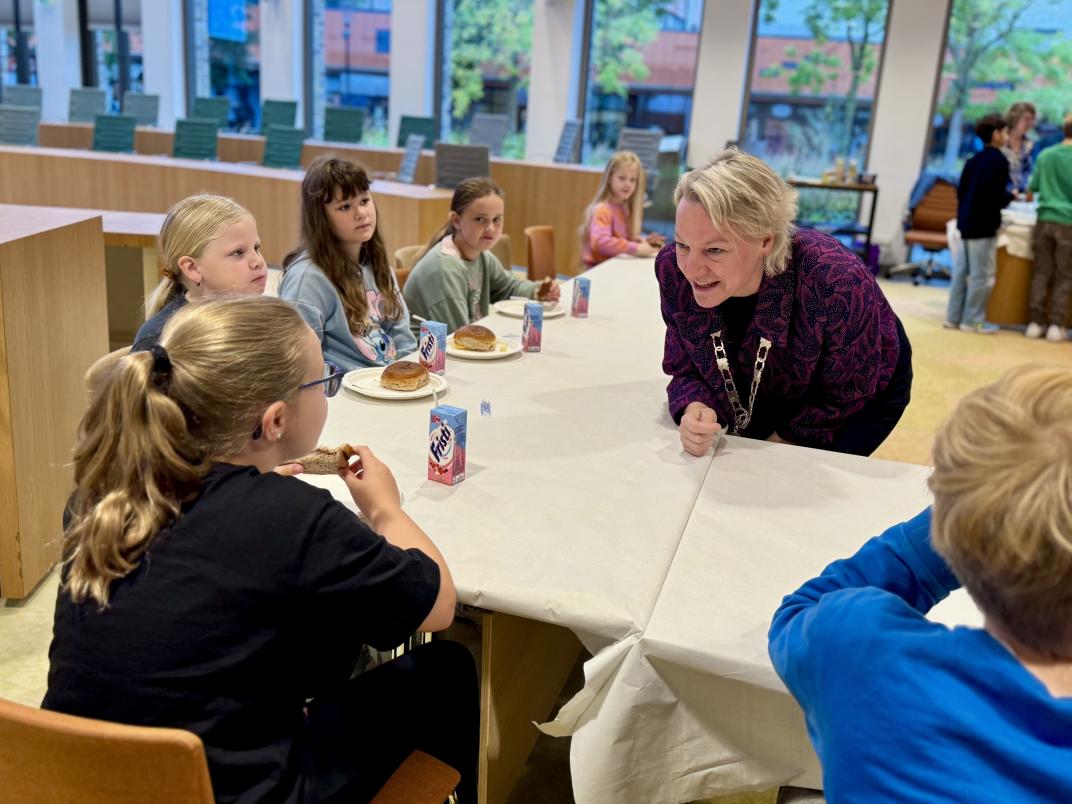 Kinderen van CBS de Wegwijzer zitten aan tafel hun ontbijt te eten. Burgemeester Erica van Lente staat bij hen en is met hen in gesprek.